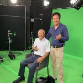 photo of Cole Kawana standing behind a seated Lawson Sakai, both are in front of a green screen with cameras in the background